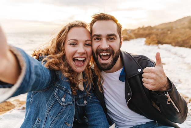 Lindo jovem casal caucasiano sorrindo e tirando uma foto de selfie enquanto caminhava à beira-mar