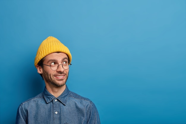 Foto grátis lindo menino milenar olha para o lado com expressão de alegria, usa chapéu amarelo e camisa jeans, tem uma ideia ou desejo interessante