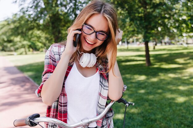 Lindo modelo feminino em fones de ouvido brancos falando ao telefone Retrato ao ar livre de garota atraente com bicicleta