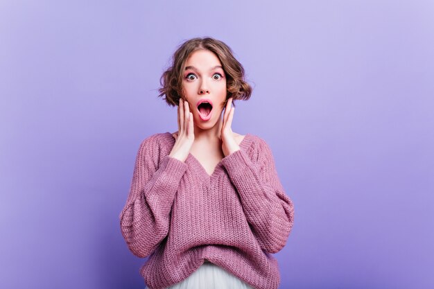 Lindo modelo feminino expressando emoções felizes durante a sessão de fotos com roupas de inverno. Retrato interno de menina bonita com um corte de cabelo elegante usa um suéter macio.