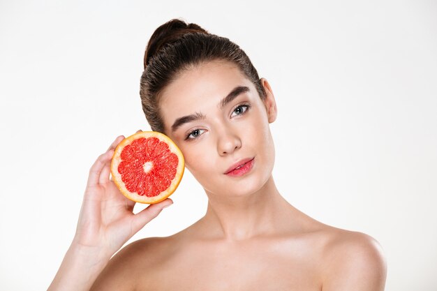 Lindo retrato de mulher bonita seminua com maquiagem natural, segurando a toranja suculenta perto do rosto e olhando