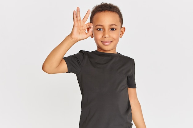 Linguagem corporal. Retrato de um garotinho de pele escura, simpático e positivo, usando uma camiseta conectando o indicador e o polegar, fazendo um gesto de aprovação, mostrando um sinal de ok, dizendo que está tudo bem