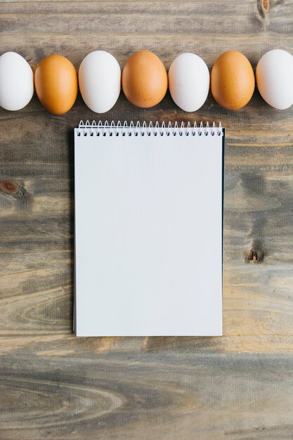 Linha de ovos marrons e brancos perto de bloco de notas em branco na mesa de madeira