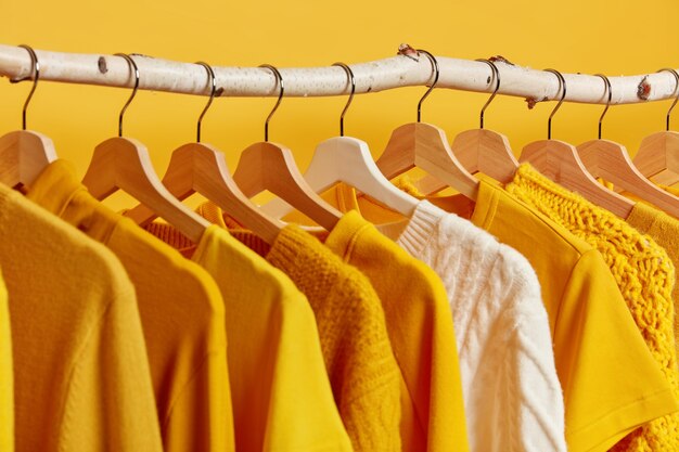 Linha de roupas da moda penduradas em um rack de madeira contra o fundo amarelo. A camisola de malha branca se destaca na coleção de inverno das roupas.
