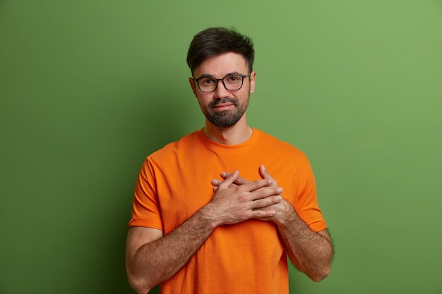 Lisonjeado satisfeito cara com a barba por fazer aperta as mãos no coração, expressa sentimentos comoventes e gratidão agradece elogios usa óculos transparentes e camiseta laranja brilhante isolada na parede verde