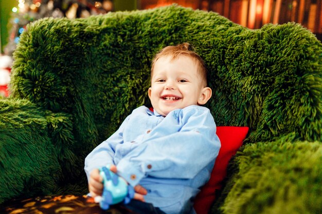 Little boy senta na poltrona verde antes de uma árvore de Natal