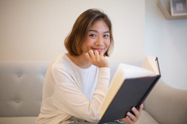 Livro de leitura de sorriso da jovem mulher no sofá em casa