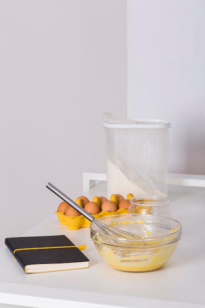 Livro de receitas; caixa de ovos; farinha e ovos batidos na mesa branca contra parede branca