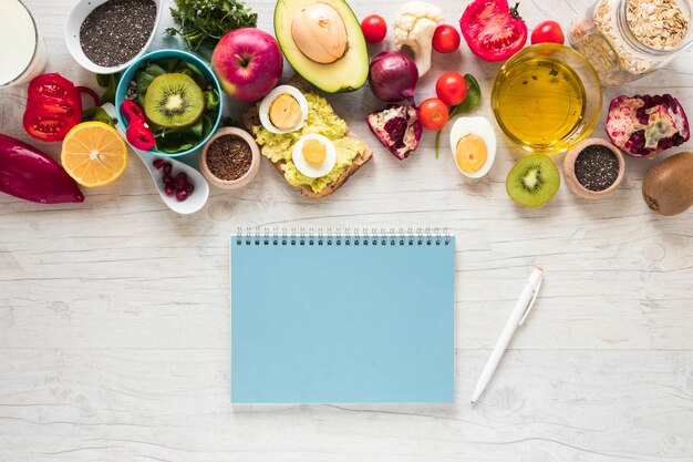 Livro espiral; caneta; frutas frescas; pão torrado; legumes e ingredientes no plano de fundo texturizado branco
