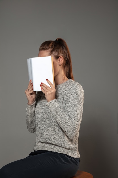 Foto grátis livro mundial feliz e dia dos direitos autorais, leia para se tornar outra pessoa - mulher cobrindo o rosto com o livro enquanto lê na parede cinza.