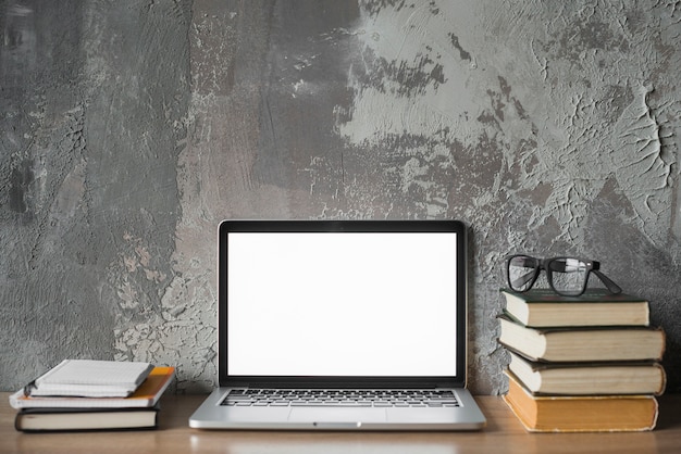 Livros empilhados; óculos e laptop com tela branca em branco na superfície de madeira