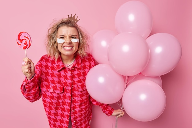 Loira bonita mulher europeia sorri o rosto aperta os dentes detém doces de caramelo e um monte de balões inflados usa coroa jaqueta xadrez manchas de beleza sob os olhos isolados sobre fundo rosa