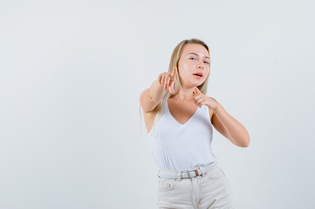 Loira de camiseta, calça apontando para a câmera e parecendo confiante