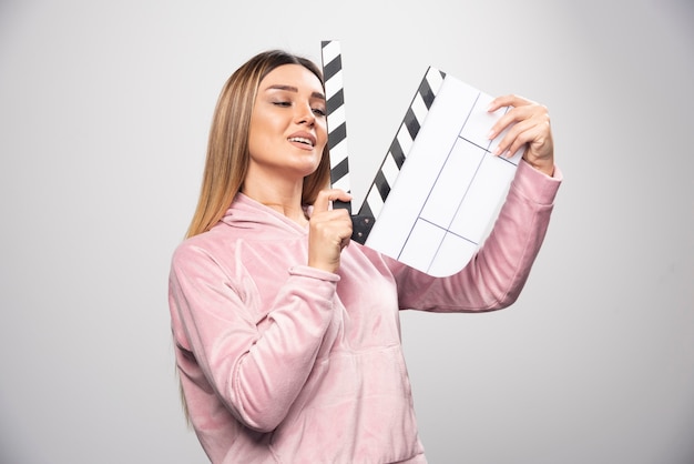 Foto grátis loira de moletom rosa segurando uma claquete em branco e dá poses positivas e divertidas.