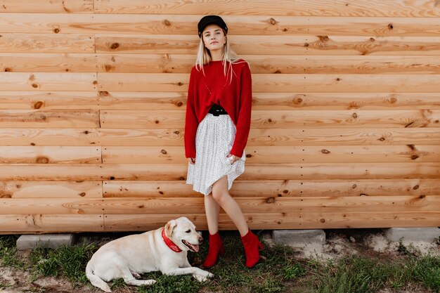 Loira deslumbrante com saia branca e pulôver vermelho, posando na parede de madeira. Linda garota se sentindo calma e bem com seu cachorro ao ar livre.