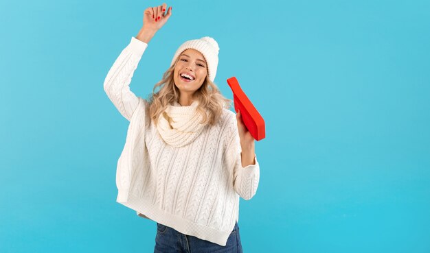 Loira elegante sorrindo linda jovem segurando um alto-falante sem fio, ouvindo música, dançando feliz vestindo um suéter branco e um chapéu de malha