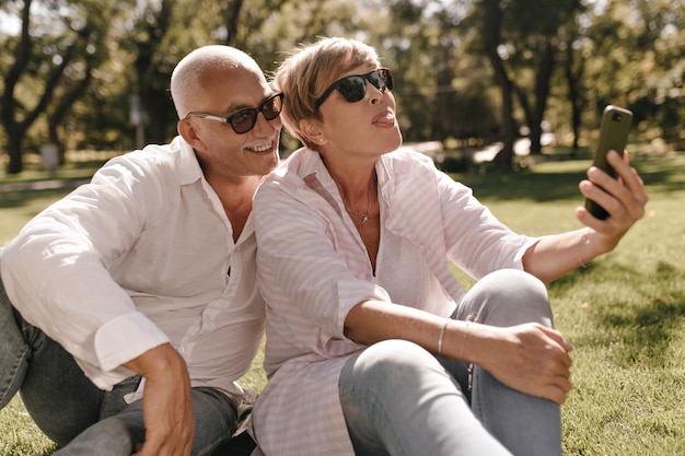 Loira engraçada em blusa listrada e jeans mostrando a língua e fazendo foto com homem sorridente em óculos de sol e camisa ao ar livre