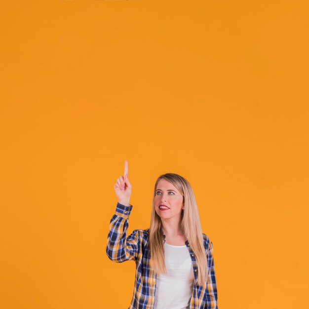 Loira jovem apontando o dedo para cima, contra um fundo laranja