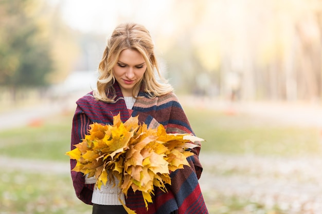 Loira sorridente oferece um buquê de folhas amarelas de outono. Bela dama em xadrez quente caminhando no parque de outono