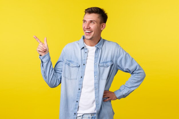 Loiro bonito e alegre, sorrindo amplamente e rindo sobre o banner promocional engraçado, apontando e olhando satisfeito no canto superior esquerdo, mostrando o anúncio do evento, fundo amarelo em pé.