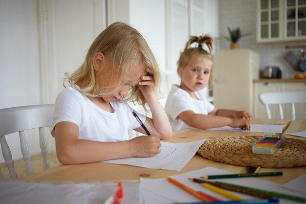 Loiro menino bonitinho fazendo lição de casa, segurando a caneta, desenhando algo na folha de papel com sua irmãzinha sentada no fundo. Duas crianças fazendo desenhos em uma mesa de madeira na cozinha