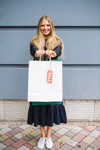 Loiro, mulher jovem, ficar, contra, parede, mostrando, bolsas para compras, com, venda, tag