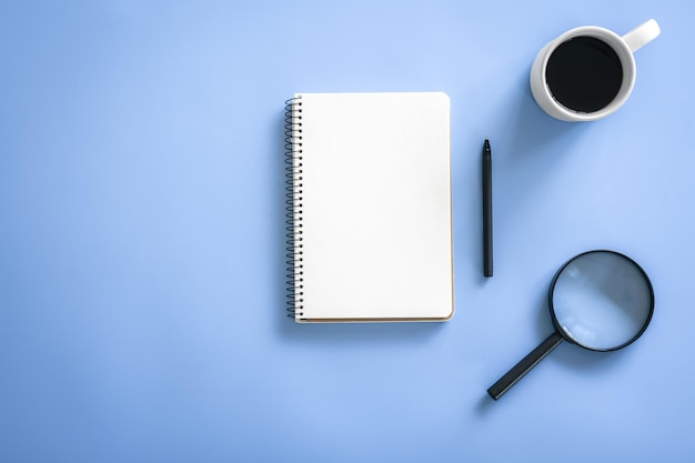 Lupa de caneta de bloco de notas e xícara de café em um plano de fundo azul
