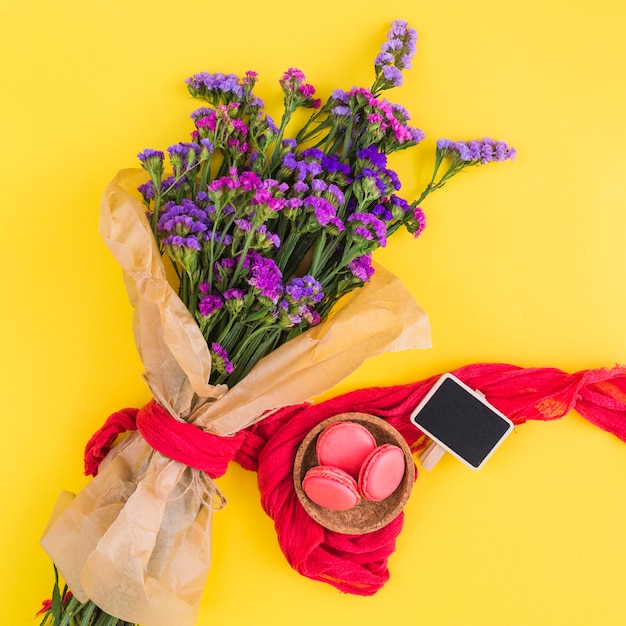 Foto grátis macaroons na tigela; cartaz em branco; buquê de flores amarrado com roupas vermelhas contra fundo amarelo