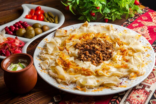 Macarrão caucasiano de guru khingal azerbaijano de vista lateral com carne picada frita e cebola com molho de creme de leite e picles em uma toalha de mesa em uma mesa de madeira escura horizontal