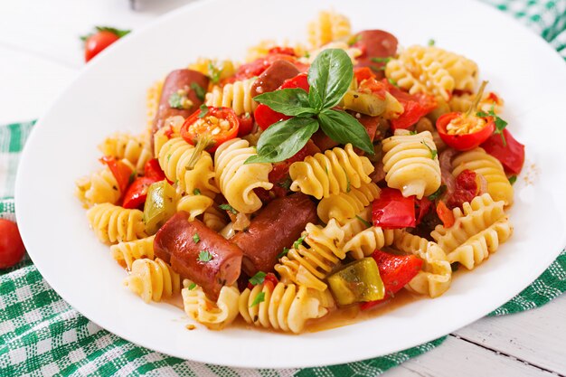 Macarrão com molho de tomate com salsicha, tomate, manjericão verde decorado em chapa branca sobre uma mesa de madeira.