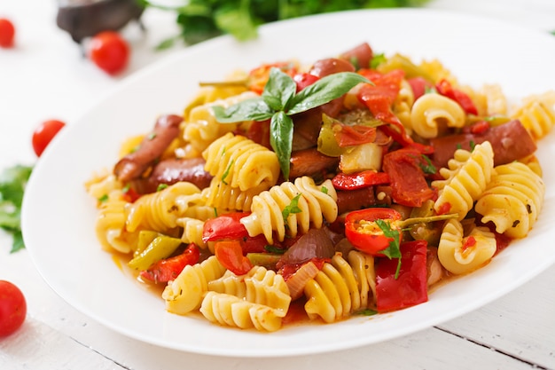 Macarrão com molho de tomate com salsicha, tomate, manjericão verde decorado em chapa branca sobre uma mesa de madeira.