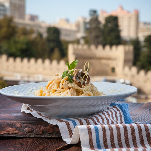 Macarrão de vista lateral com queijo e cogumelos em um prato branco sobre uma mesa de madeira escura com vista cidade