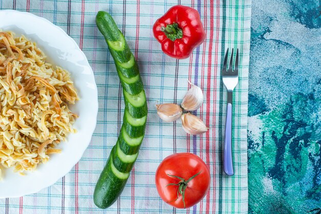 Macarrão em um prato ao lado de vários vegetais e garfo em uma toalha de chá, na mesa azul.