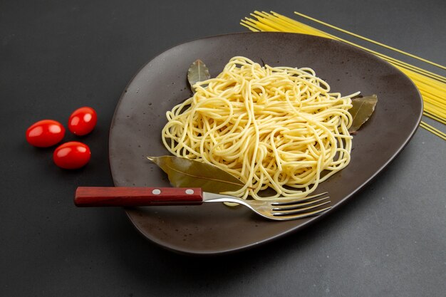 Macarrão espaguete com folhas de louro, garfo no prato, tomate cereja, macarrão espaguete cru em fundo preto