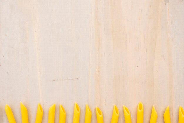 Macarrão penne amarelo cru, disposta em linha no plano de fundo texturizado de madeira