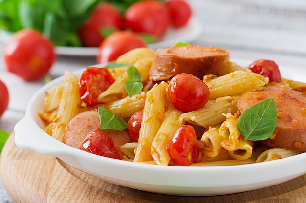 Macarrão penne com molho de tomate com linguiça, tomate, manjericão verde, decorado em uma frigideira sobre uma mesa de madeira