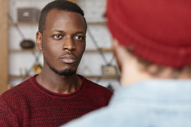 Macho americano africano jovem bonito vestindo blusa casual falando com seu amigo caucasiano irreconhecível, ouvindo-o com interesse e atenção. Foco seletivo no rosto do homem negro