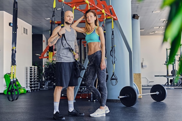 Macho atlético de cabeça raspada e fêmea morena magra posando perto de tiras trx fica em um clube de ginástica.