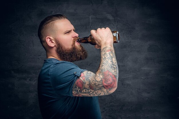 Macho barbudo brutal com braço tatuado bebe uma cerveja de uma garrafa.