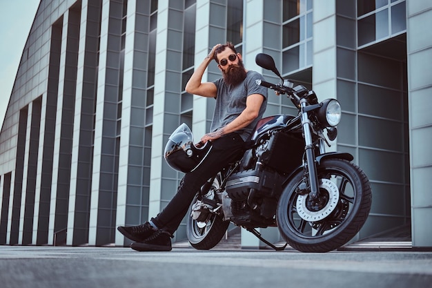 Foto grátis macho barbudo brutal de óculos vestido com uma camiseta cinza e calça preta ajeita o cabelo e segura um capacete sentado em sua motocicleta retrô feita sob medida contra um arranha-céu.