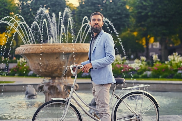 Macho barbudo de luxo em óculos de sol em uma bicicleta sobre fundo de fonte da cidade.