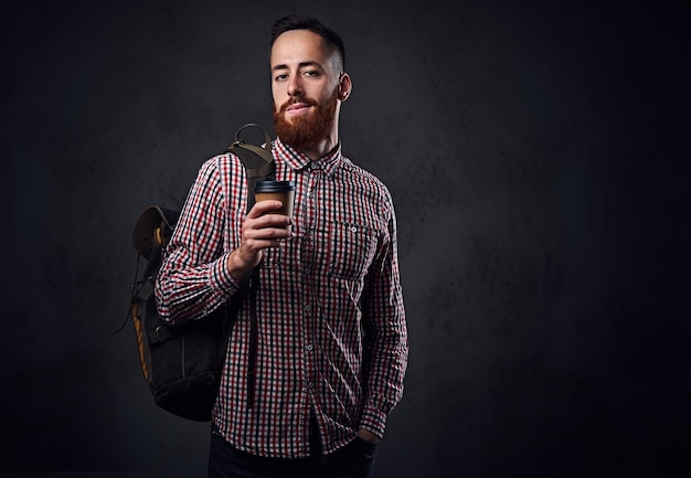 Macho barbudo ruivo vestido com uma camisa de lã segura uma mochila e tira a xícara de café.