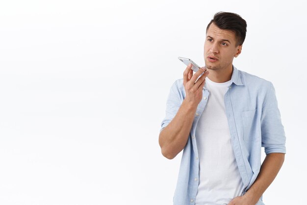 Macho bonito de aparência séria na camisa grava mensagem de voz ou memorando no aplicativo de smartphone segurando o celular perto dos lábios perto da dinâmica falando com o fundo branco do assistente de pessoa