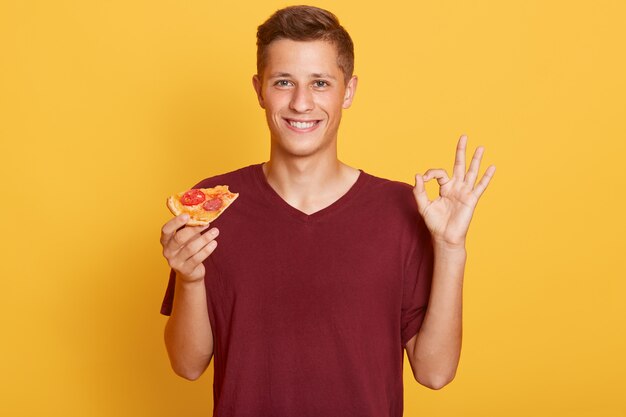 Macho bonito veste camiseta marrom casual, segurando a fatia de pizza nas mãos