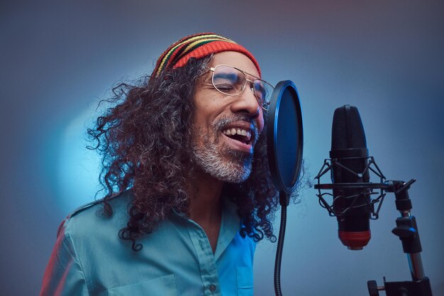 Macho cantor rastafari africano vestindo uma camisa azul e gorro escrevendo música emocionalmente no estúdio de gravação. Isolado em um fundo azul.