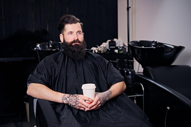 Macho de hipster barbudo a todo vapor bebe café antes do penteado no salão de cabeleireiro.