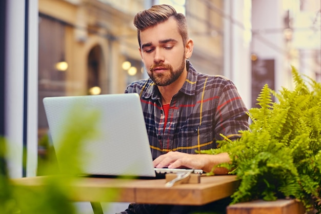 Macho de olhos azuis e barbudo usando um laptop em um café na rua.