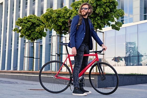 Macho hipster barbudo com mochila senta-se na bicicleta fixa vermelha em um parque.