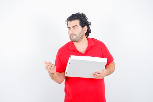 Macho maduro em uma camiseta vermelha segurando uma caixa de pizza enquanto olha para longe e olhando pensativo, vista frontal