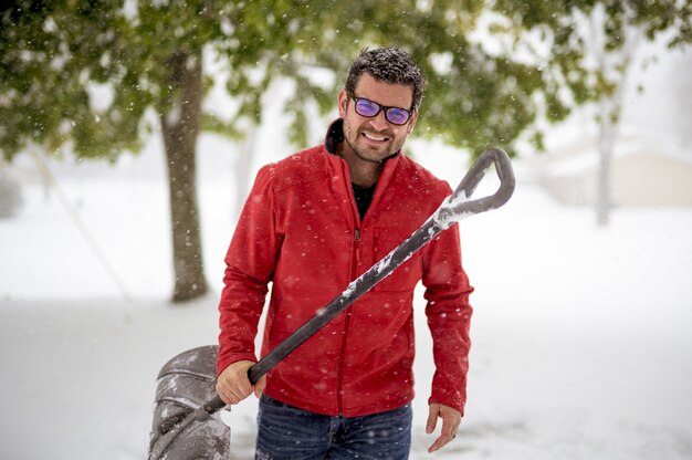 Macho segurando uma pá de neve e vestindo uma jaqueta vermelha enquanto sorrindo